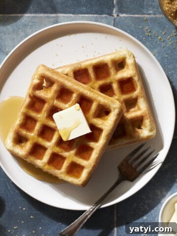 Cropped image of two Sourdough Discard Waffles on a white plate with a pad of butter and maple syrup.