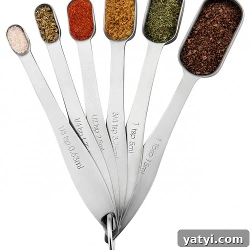 A set of six metal measuring spoons, each filled with a different spice or herb, fanned out in a semi-circle and connected by a metal ring.