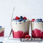 Side view of a glass cup with a spoon leaning up against the side and layers of chia seed pudding - raspberry, vanilla, and fresh blueberries on top. Surrounded by fresh berries on a white background