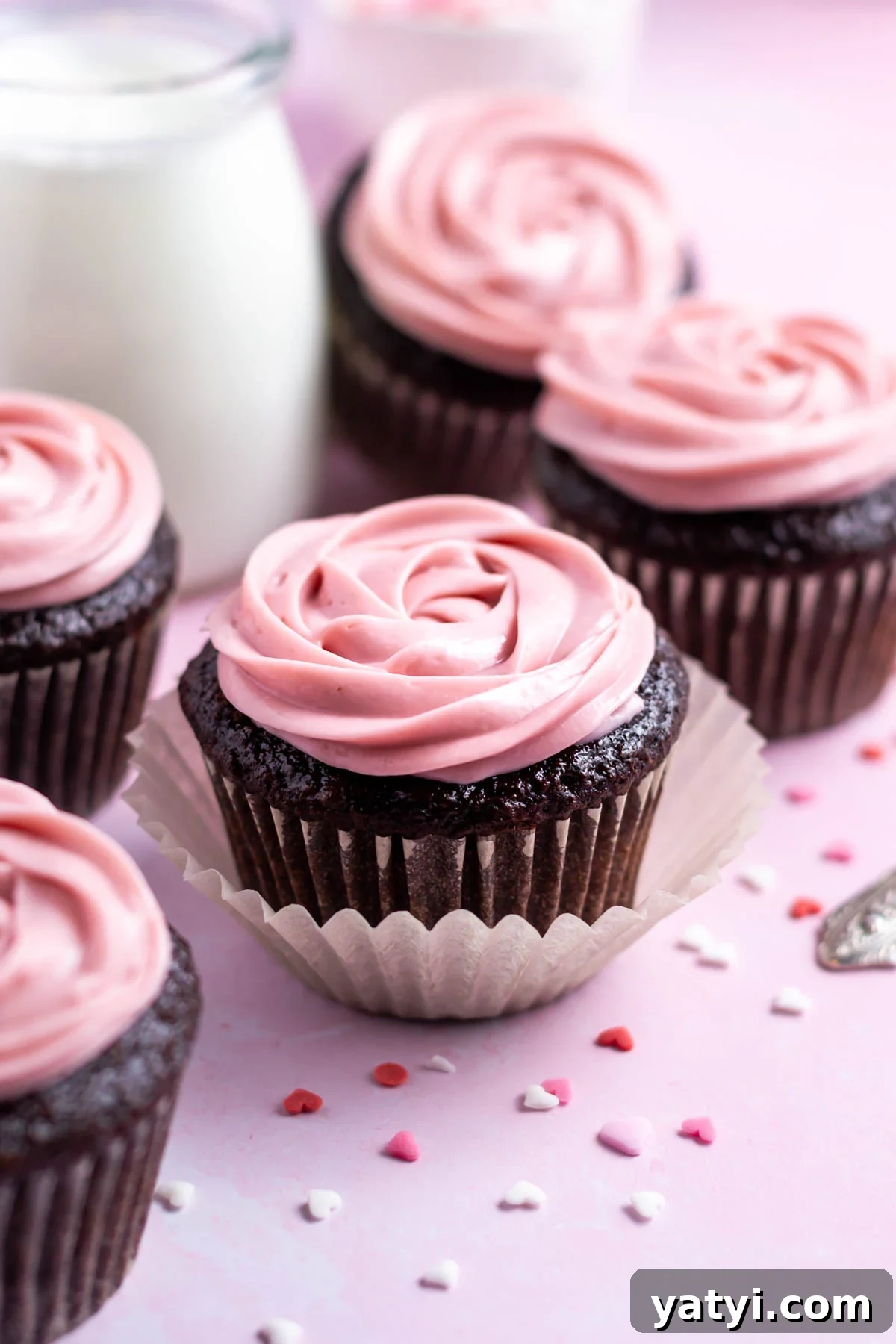 Moist Chocolate Cupcakes with Raspberry Filling and Ganache 2 chocolate raspberry cupcakes with heart shaped sprinkles and a glass of milk in the background