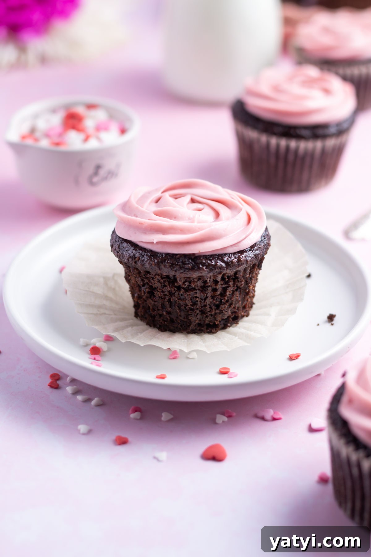 Moist Chocolate Cupcakes with Raspberry Filling and Ganache 16 chocolate cupcake with the wrapper missing on a plate with a bowl of sprinkles and milk in the background