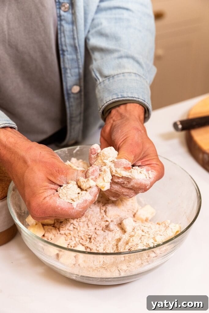 Chewy Cinnamon Apple Pie Bars: Easy Recipe for Fall Baking 7 Two hands pressing butter with flour mixture in large glass bowl on white surface.