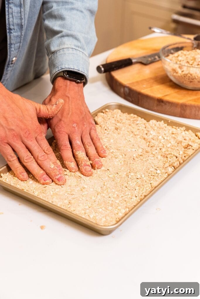 Chewy Cinnamon Apple Pie Bars: Easy Recipe for Fall Baking 9 Two hands pressing crust for apple pie bars into a large gold baking pan sitting on white surface.