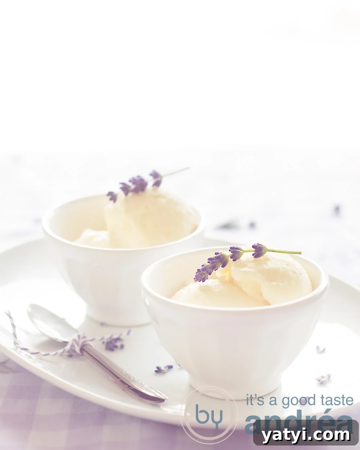 Two white bowls filled with lavender flavored ice cream and a fresh sprig of lavender. They are on a white plate with two spoons on top. A pink white background and a white background.