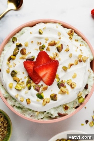Overhead view of pistachio fluff in a pink ceramic bowl with strawberry slices, pistachios, and whipped cream garnish, surrounded by ingredient bowls on marble surface.