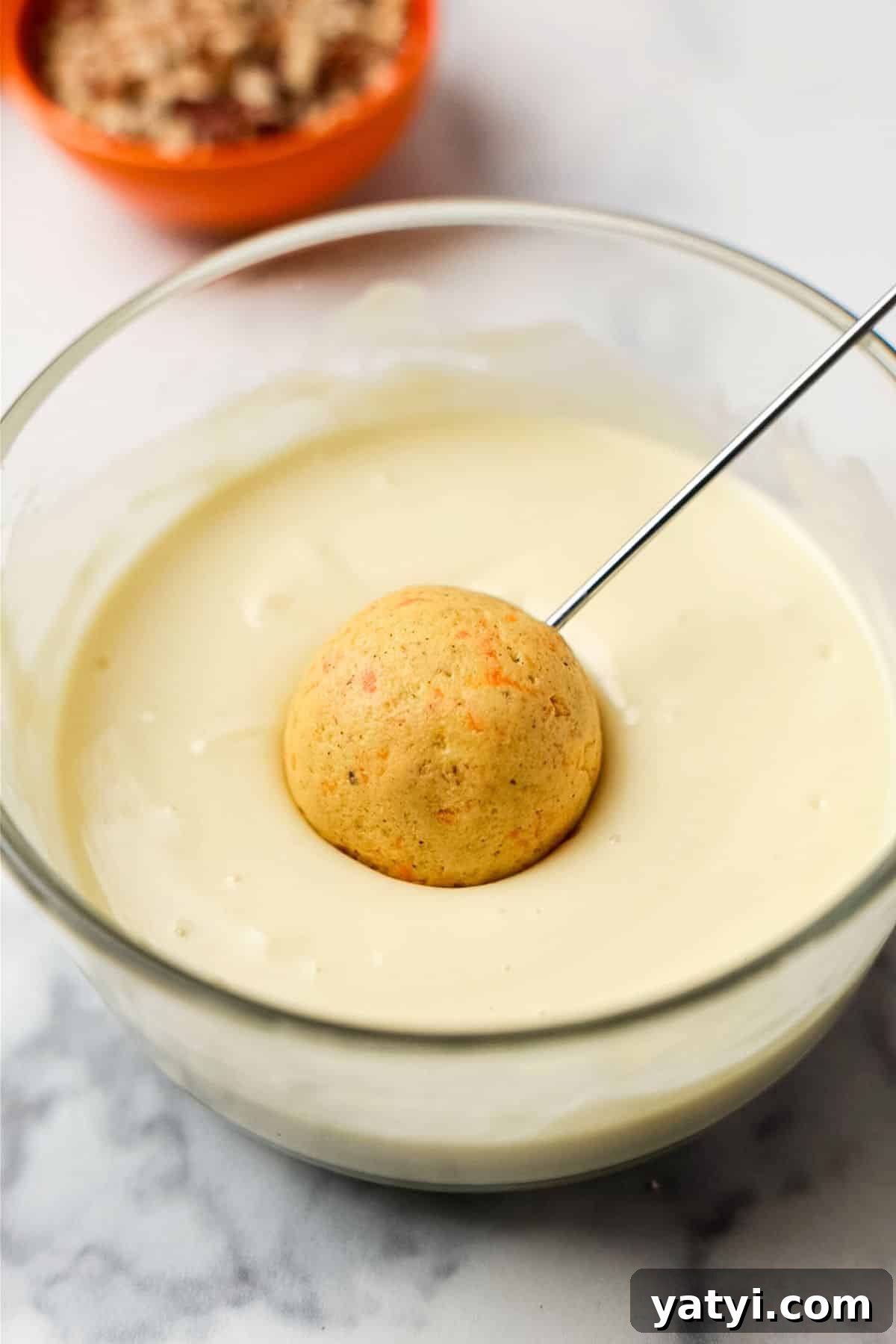 Keto Carrot Cake Protein Bites Recipe for Low-Carb Snacks 8 A carrot cake bite being lowered into melted white chocolate in a glass bowl, with chopped pecans in the background