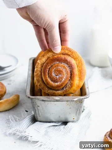 Hand pulling a skillingsboller out of a silver loaf pan with plates, a glass of milk and other skillingsboller around it.