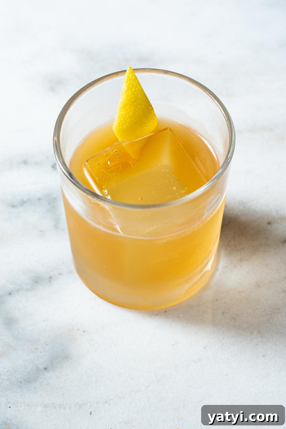 Gold Rush Whiskey Cocktail Recipe: Classic Honey-Lemon Sour 2 A golden Gold Rush cocktail on a marble surface, served with a giant clear ice cube and lemon peel garnish.