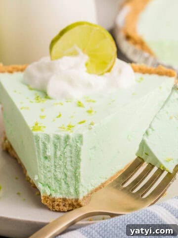 Close-up of a slice of key lime Jello pie with a bite on a fork beside it. The pie is topped with whipped cream, lime zest, and a lime slice, showing its airy and creamy whipped texture.