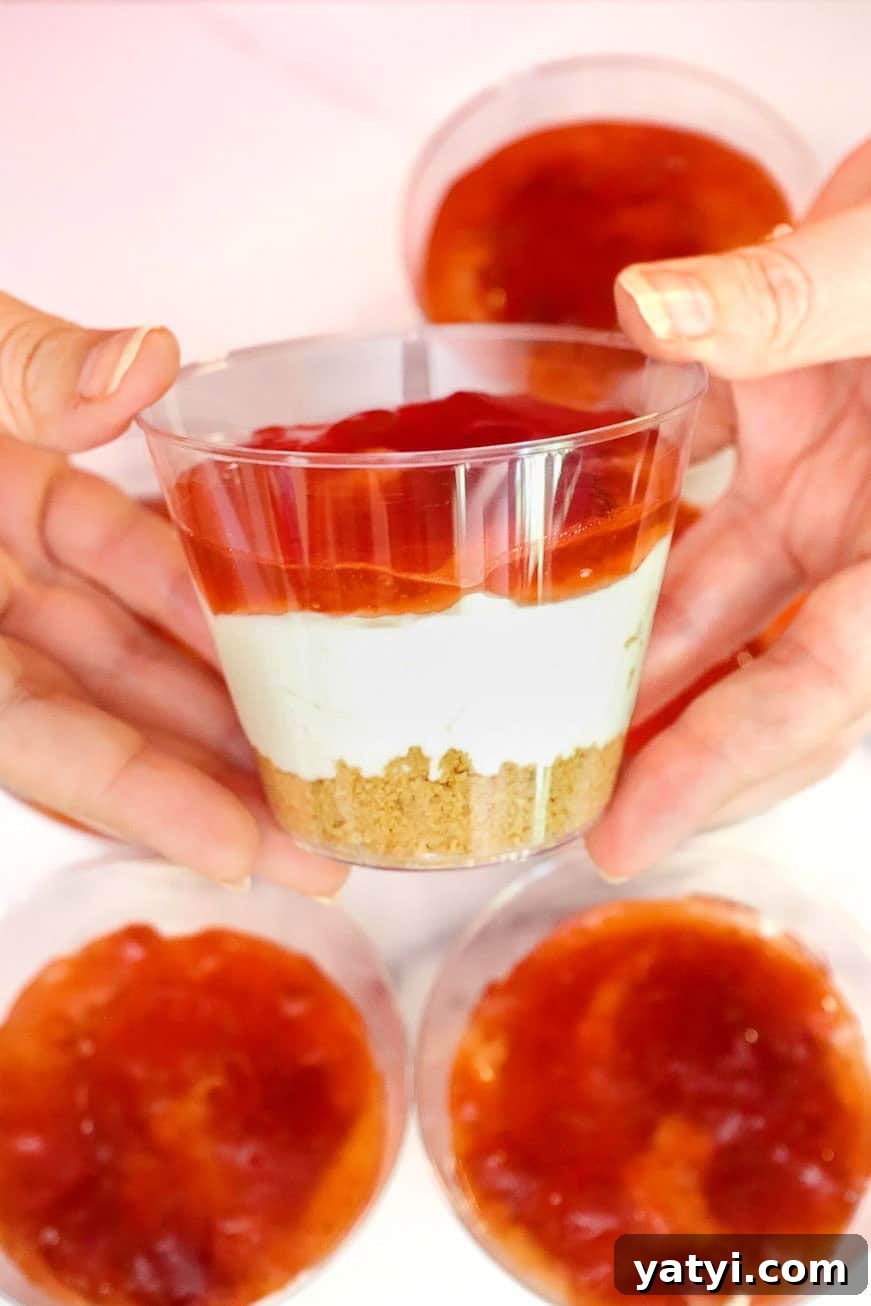 No-Bake Strawberry Cheesecake Cups with Graham Cracker Crust 8 Hands holding a cup layered with graham cracker crust, cheesecake filling, and strawberry jam.