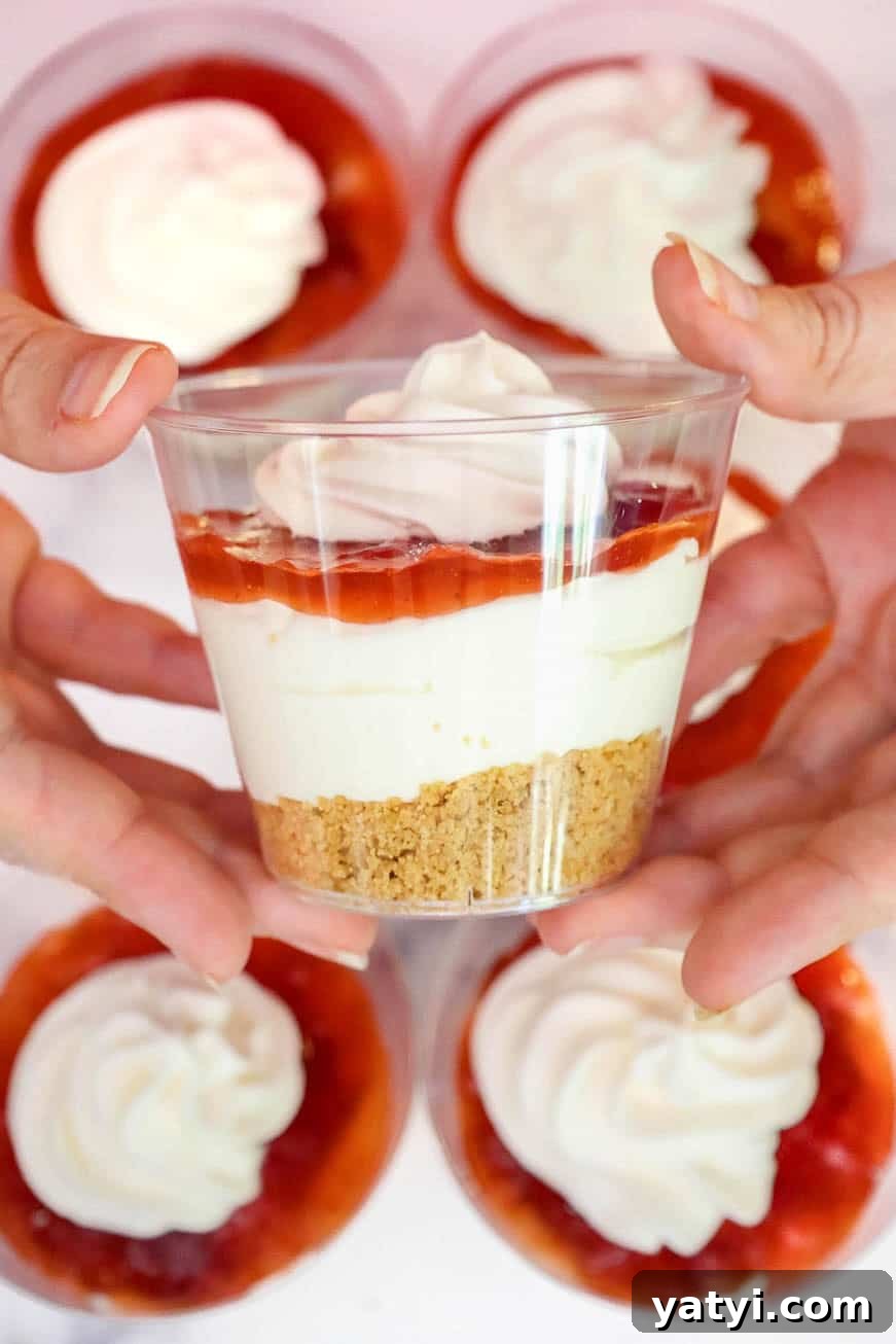 No-Bake Strawberry Cheesecake Cups with Graham Cracker Crust 9 Hands holding a finished cheesecake cup topped with whipped cream. Other cups with whipped topping are in the background.