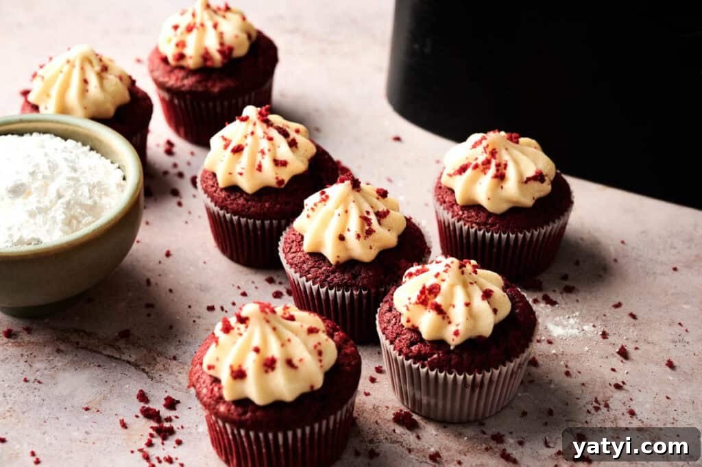 Six Air Fryer Red Velvet Cupcakes with cream cheese frosting, topped with red crumbs, are arranged on a light surface next to a bowl of powdered sugar.