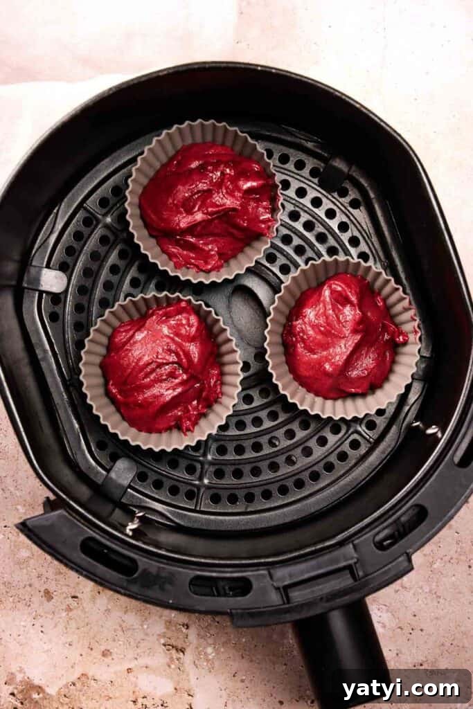 Three silicone cupcake liners filled with red batter are arranged inside an air fryer basket, ready to become delicious Air Fryer Red Velvet Cupcakes.