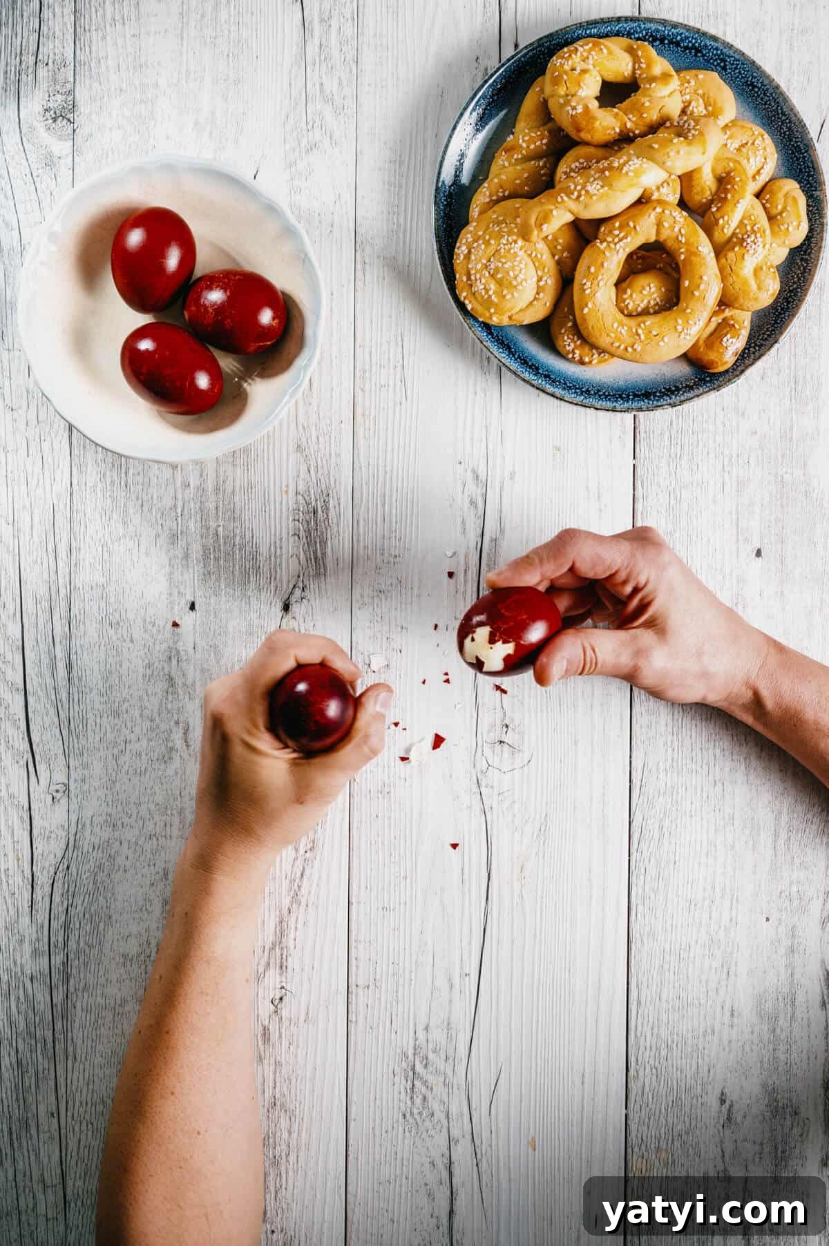 Greek Easter Traditions: Customs, Foods, and Celebrations Explained 2 two hands cracking Greek Easter eggs with cookies on a white wooden table.