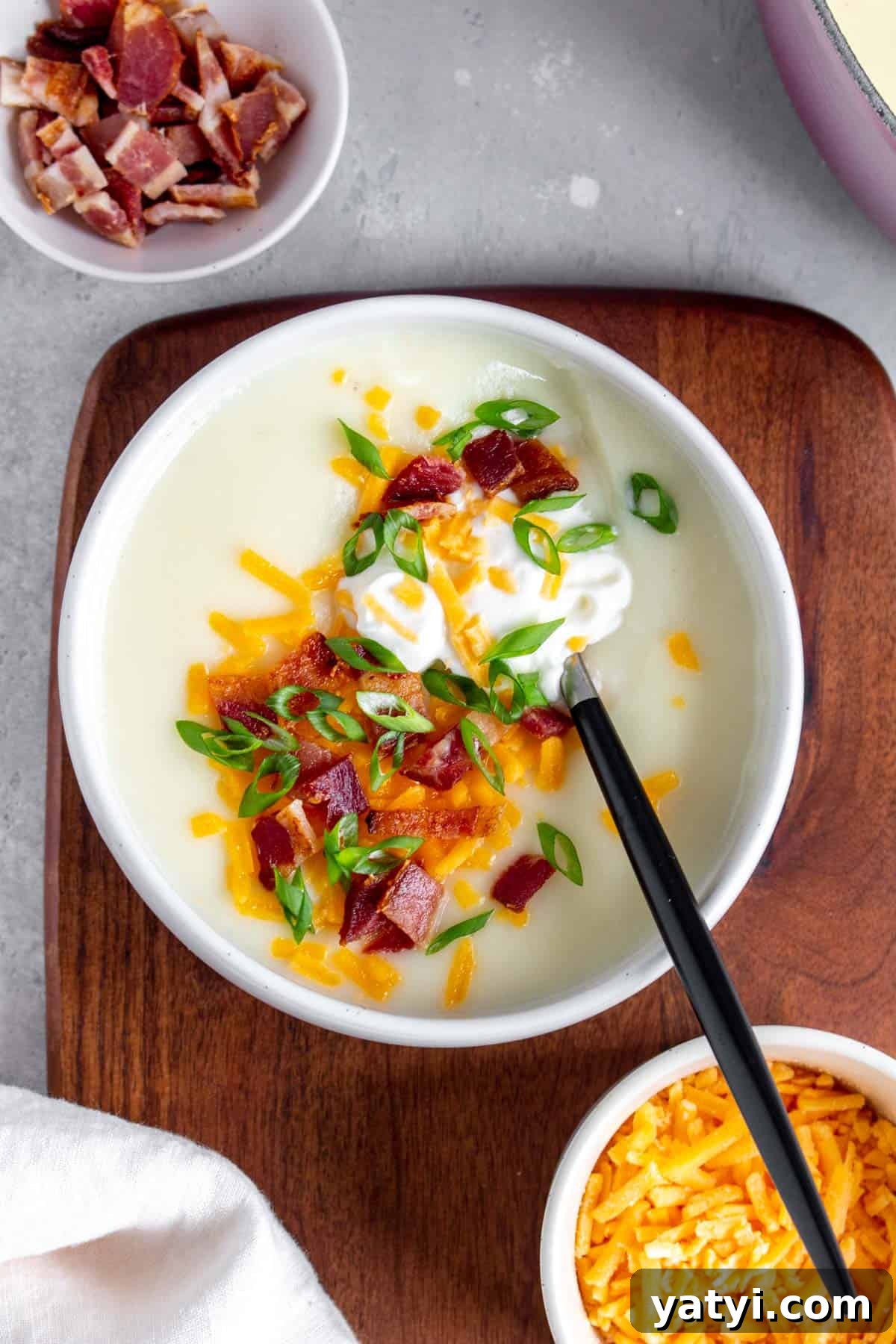 Creamy 4-Ingredient Potato Soup Recipe 2 A bowl of potato soup with sour cream, shredded cheese, bacon, and green onions with a spoon inside.