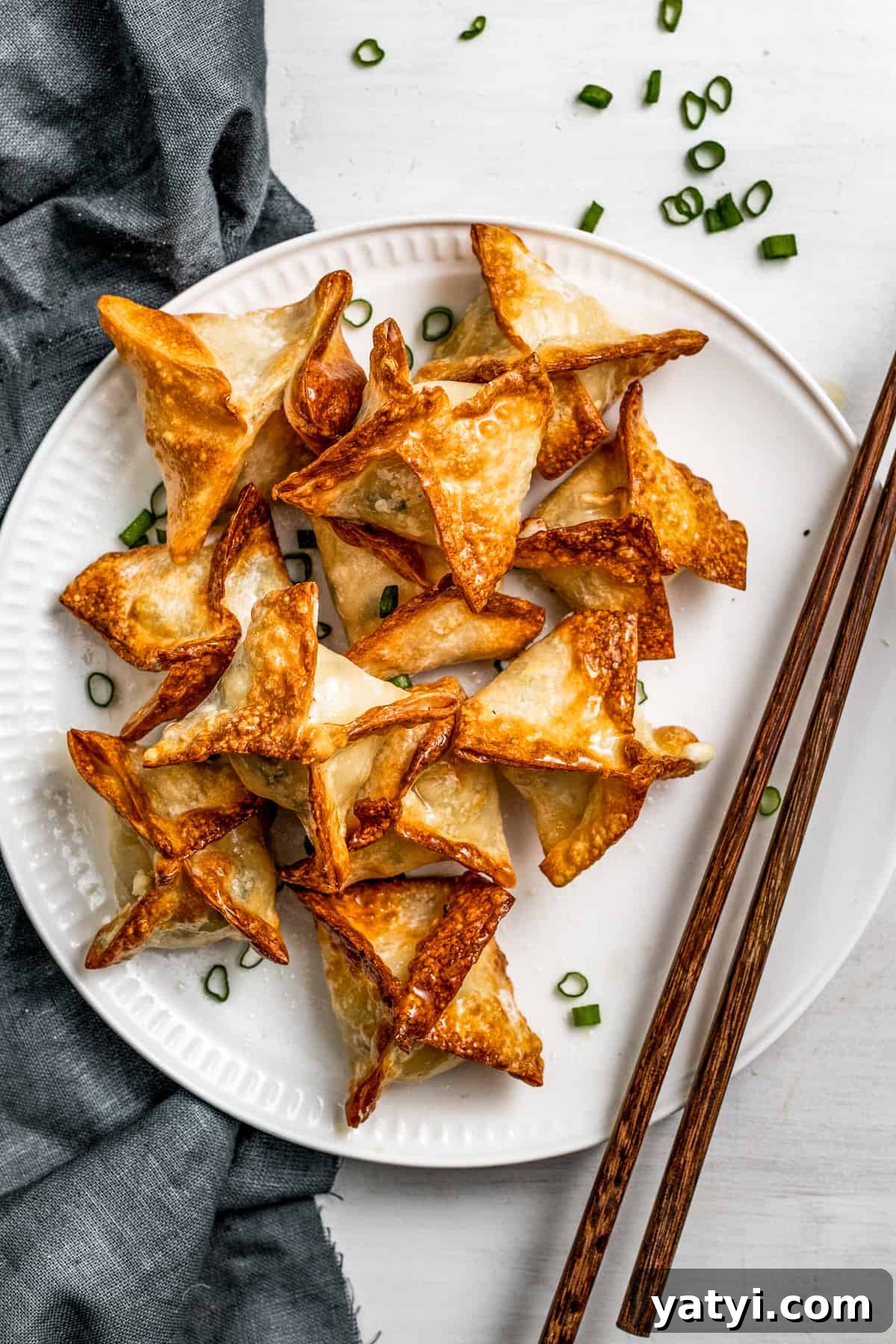 Air fryer crab rangoon are crispy and golden. Crab rangoon on a white plate garnished with green onions and a pair of chopsticks placed next to the rangoons.