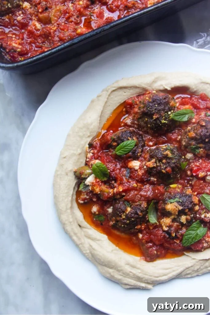 Meatballs in tomato sauce on top of creamy hummus on a white plate, with dish of tomatoes and meatballs in the background.