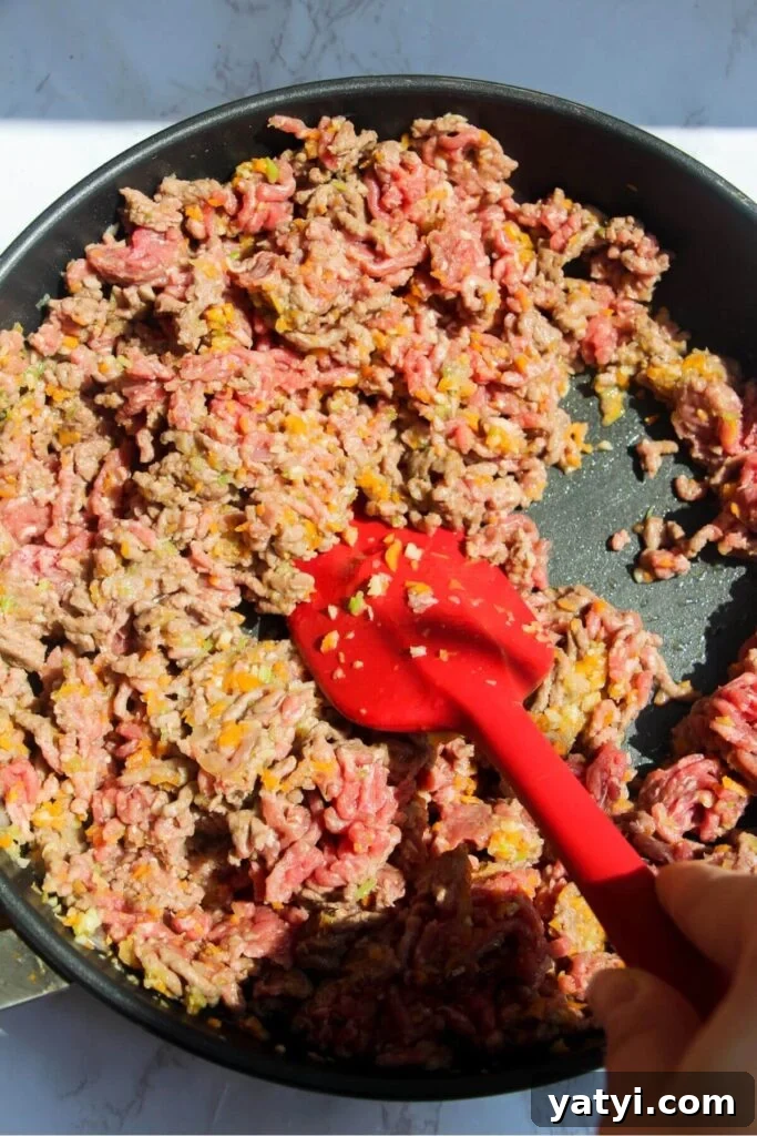 Red spatula stirring ground beef and browning it in a large pan.