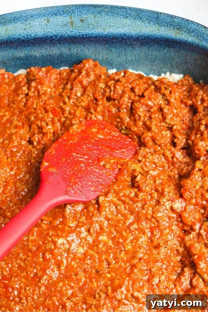 Red spatula pressing bolognese into a blue oven dish.