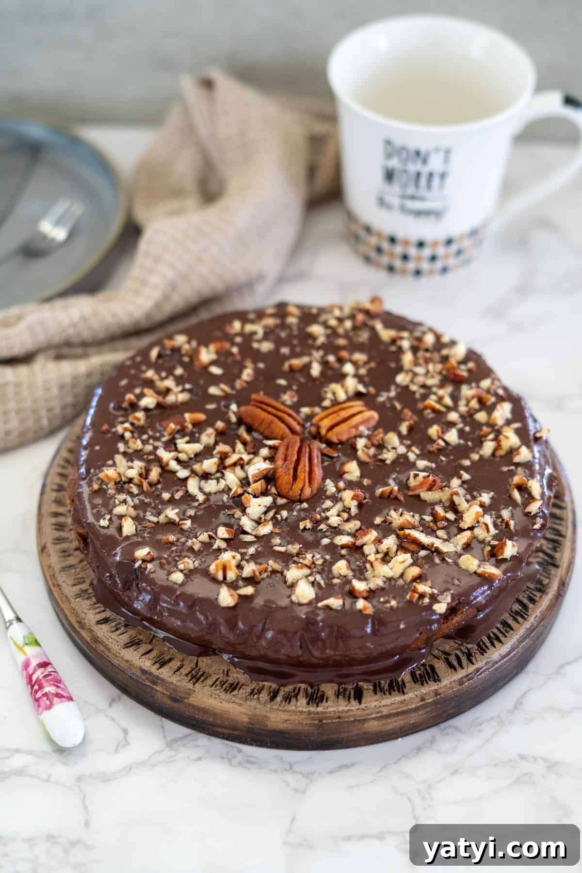 Keto Chocolate Pecan Torte Recipe – Low-Carb Dessert Delight 2 A decadent pecan torte, topped with chopped nuts and rich chocolate, rests on a wooden plate. It's accompanied by a mug and a knife elegantly placed on the cool marble surface.