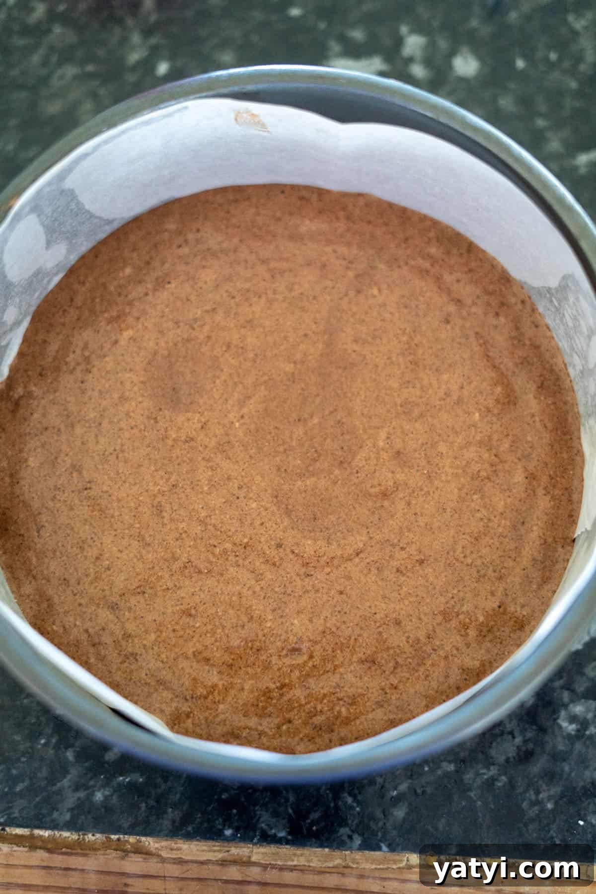 Keto Chocolate Pecan Torte Recipe – Low-Carb Dessert Delight 10 Brown batter for a pecan torte rests in a round baking tin lined with parchment paper on a dark countertop.
