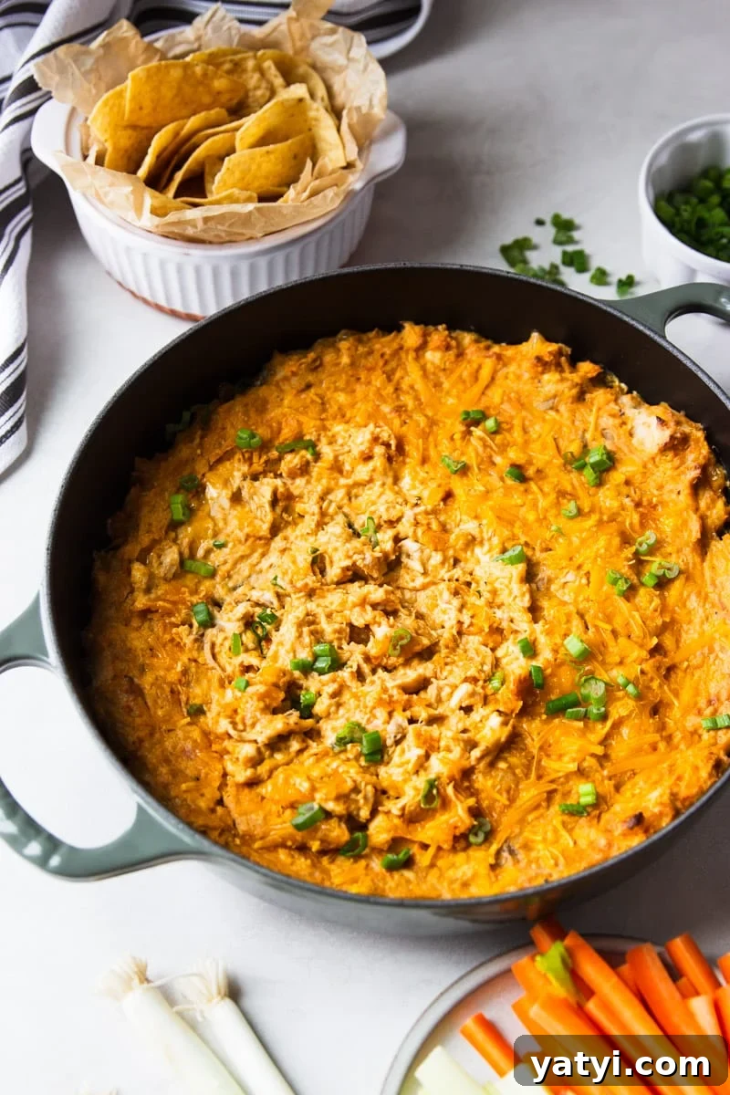 Creamy Buffalo Chicken Dip Recipe for Game Day or Parties 3 Green onions in a white bowl, corn chips in a white serving bowl, white and black dish towel, baked buffalo chicken dip in a skillet, carrots and celery.