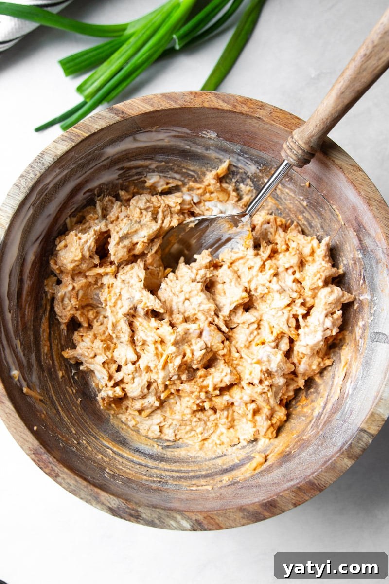 Creamy Buffalo Chicken Dip Recipe for Game Day or Parties 5 Green onions, spoon with wooden handle, shredded chicken mixed with cheese in a wooden bowl.