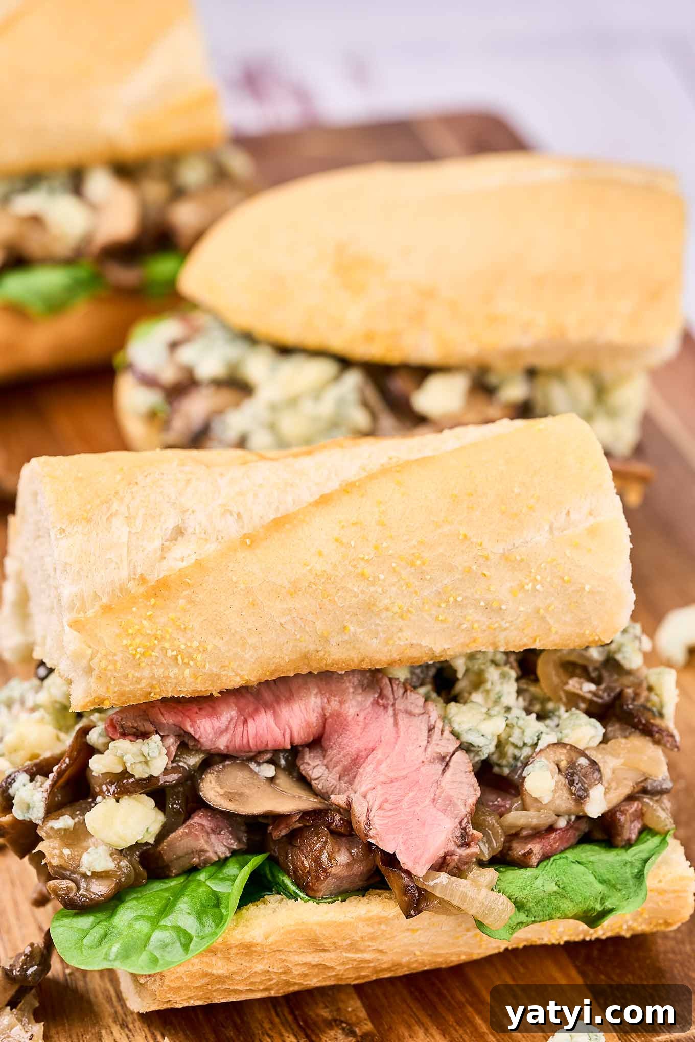 Ribeye Steak Sandwich with Creamy Blue Cheese Sauce 2 A wooden cutting board with three portions of grilled ribeye steak sandwich with mushrooms, onions, fresh spinach, and blue cheese on a French baguette.