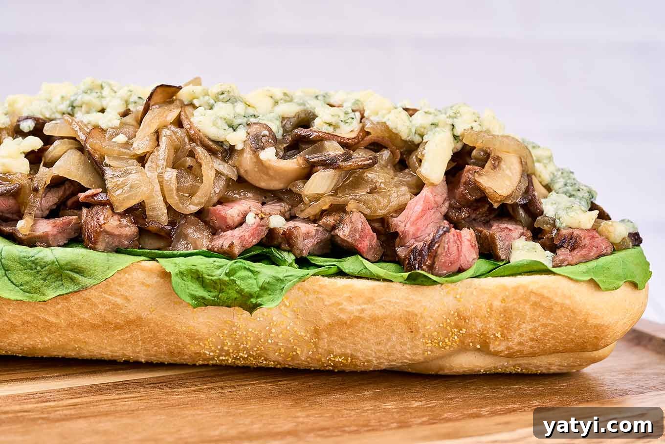 Ribeye Steak Sandwich with Creamy Blue Cheese Sauce 5 A split baguette filled with grilled ribeye, sautéed onions and mushrooms, spinach, and blue cheese on a cutting board.