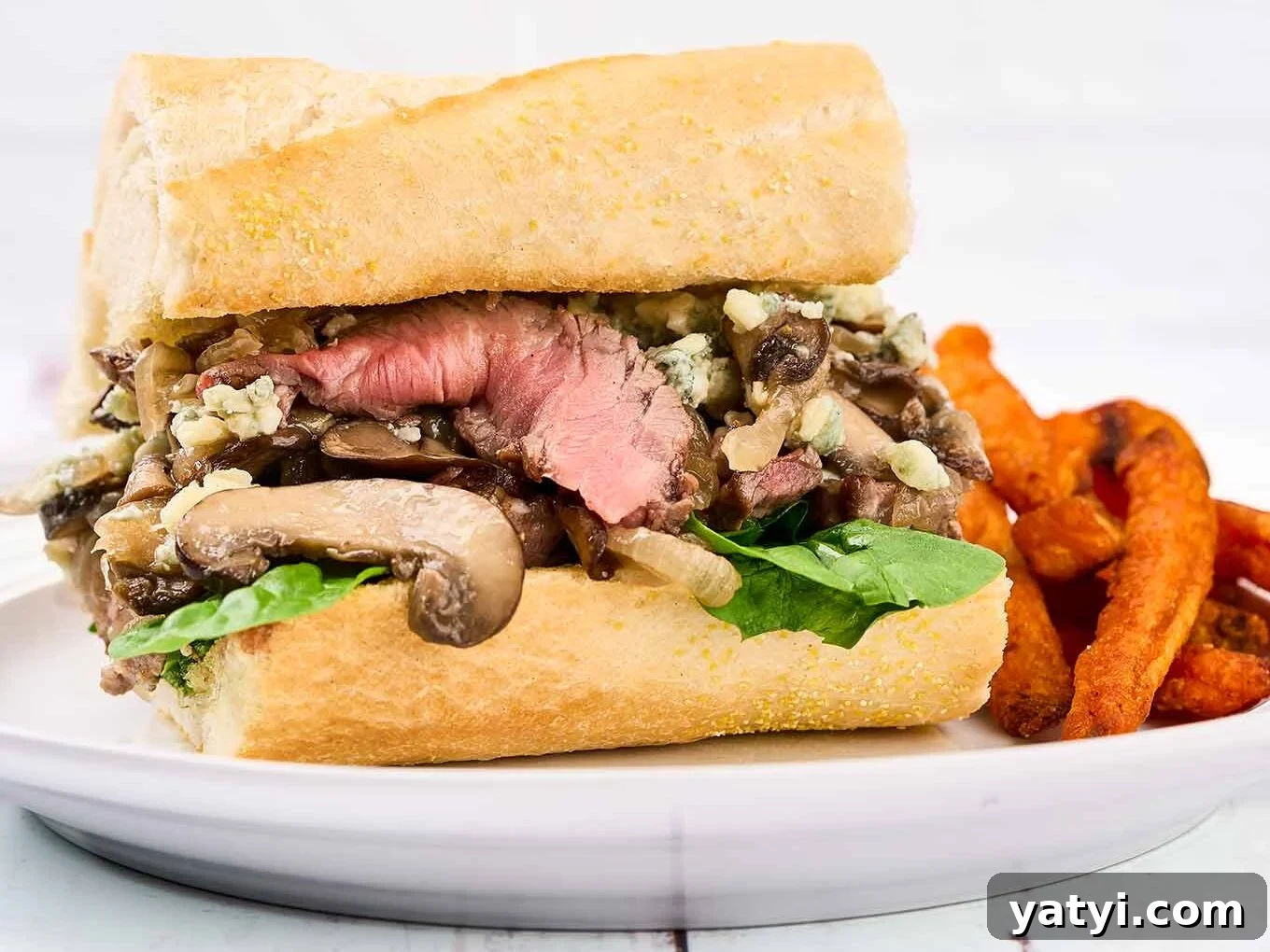Ribeye Steak Sandwich with Creamy Blue Cheese Sauce 6 A single portion of a grilled ribeye steak sandwich with blue cheese on a white oval plate with sweet potato fries on the side.