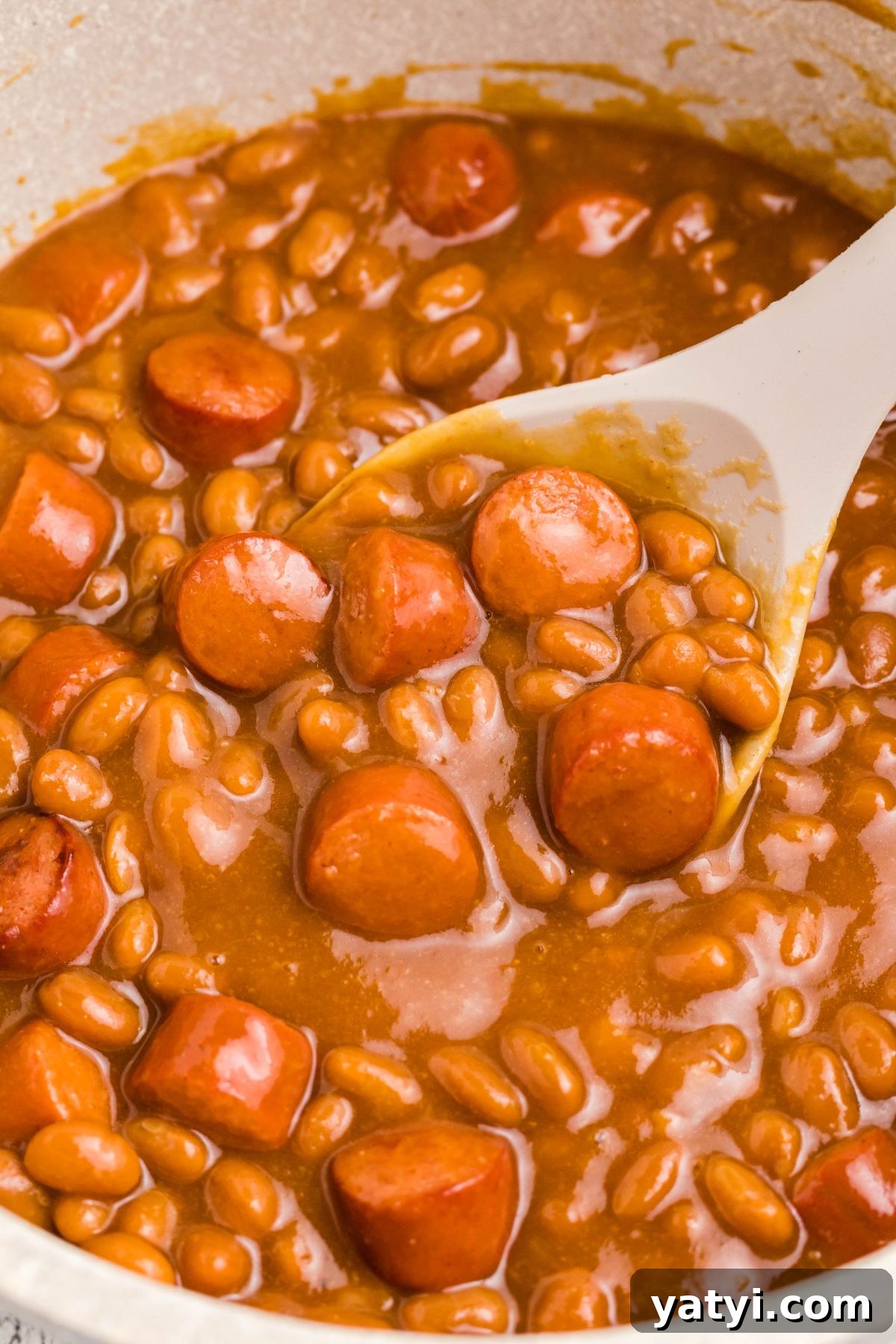 Classic Beanie Weenies Recipe: Hearty Sausage and Beans Meal 7 Close up photo of beans and weenies in a saucepan with a large spoon in the center.