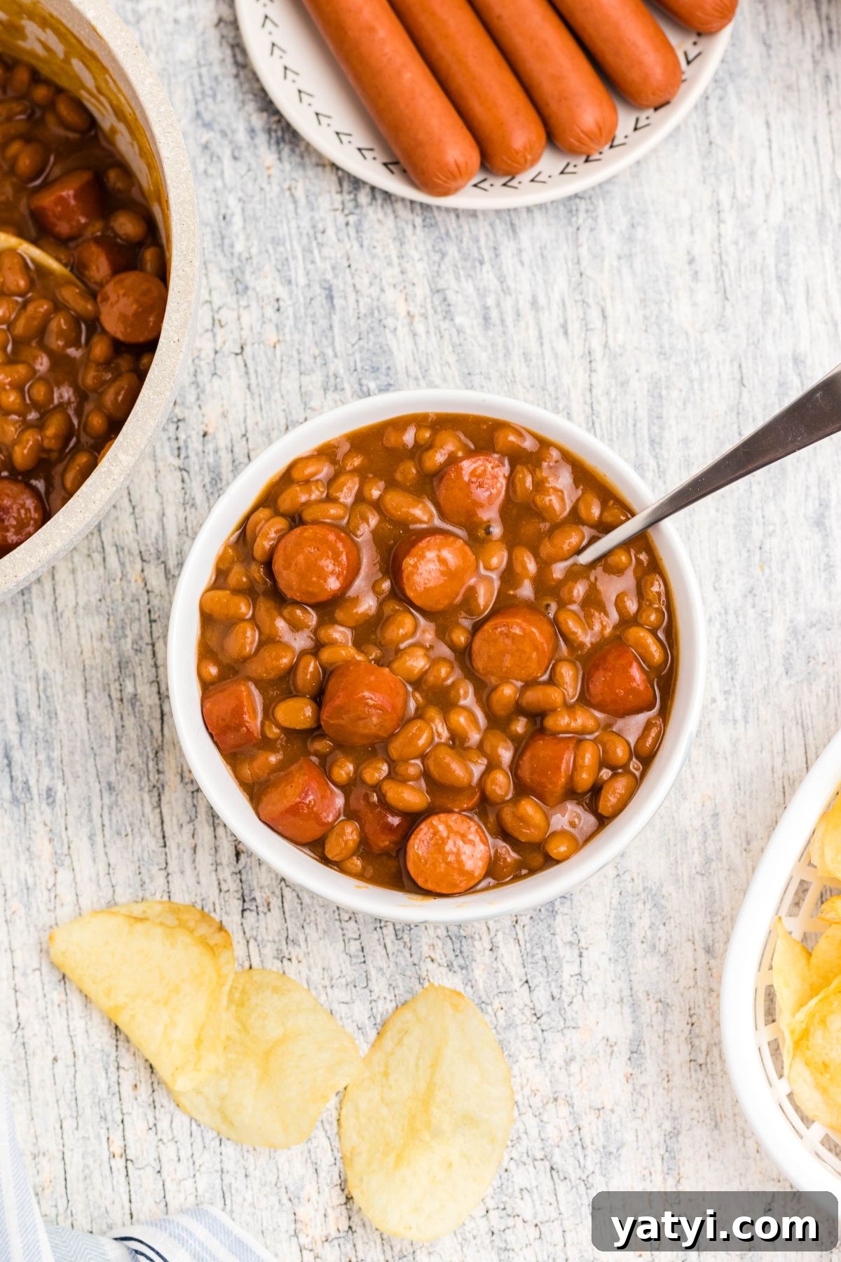 Classic Beanie Weenies Recipe: Hearty Sausage and Beans Meal 8 Cut up hot dogs, mixed with beans, in a white bowl with potato chips scattered around the dish.