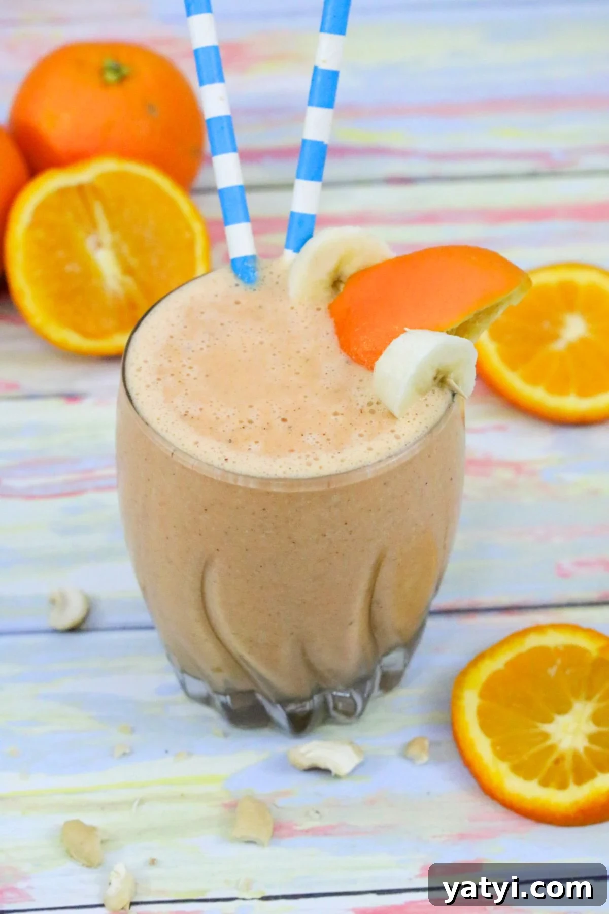 Orange and carrot smoothie in a glass with banana and orange slice on the side and blue straws in it.
