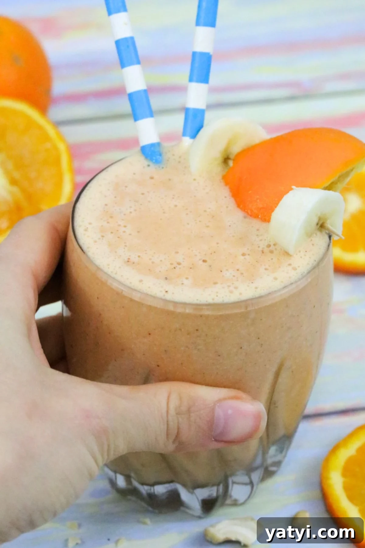 Orange and carrot smoothie in a glass with banana and orange slice on the side and blue straws in it and a hand holding the glass.