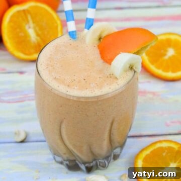 Orange and carrot smoothie in a glass with banana and orange slice on the side and blue straws in it.