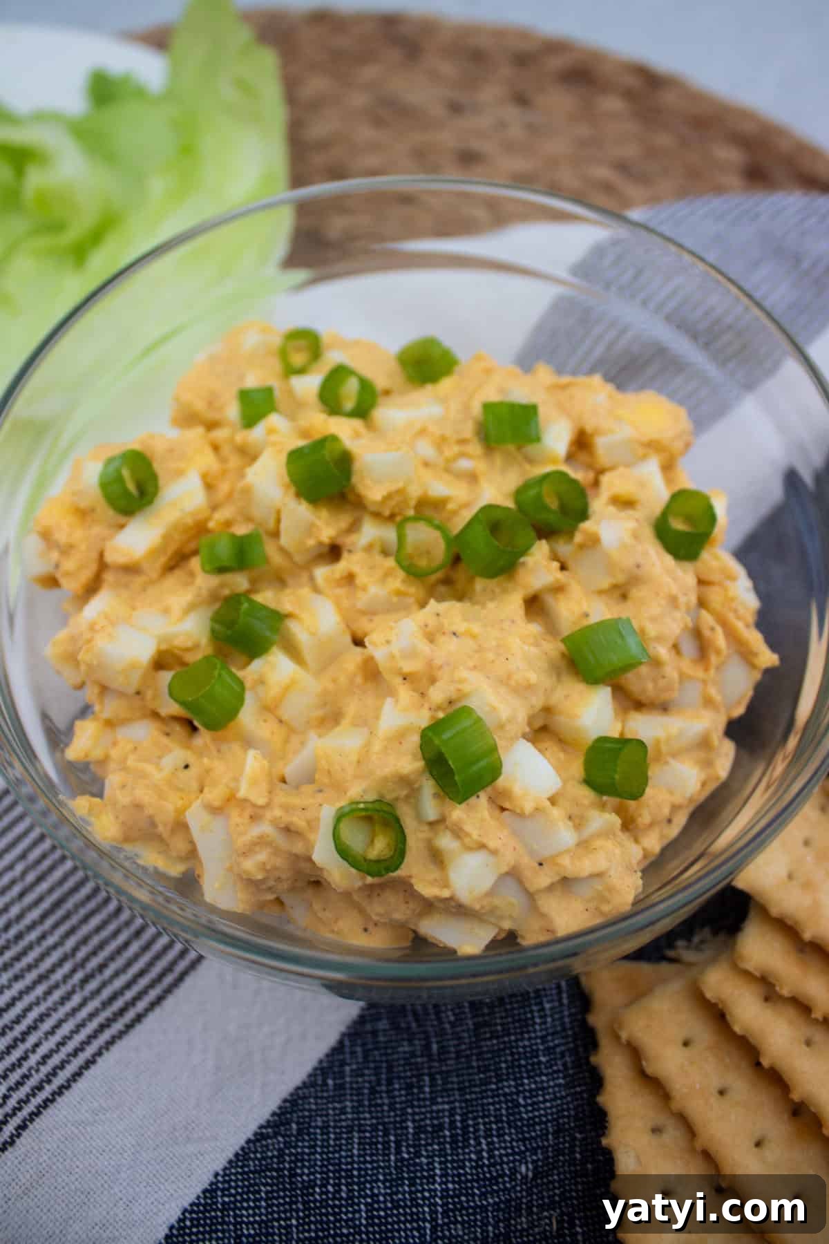 Creamy Egg Salad Without Mayo: Tangy Avocado & Yogurt Recipe 2 A glass bowl filled with egg salad topped with sliced green onions, placed on a striped cloth next to lettuce leaves and square crackers.
