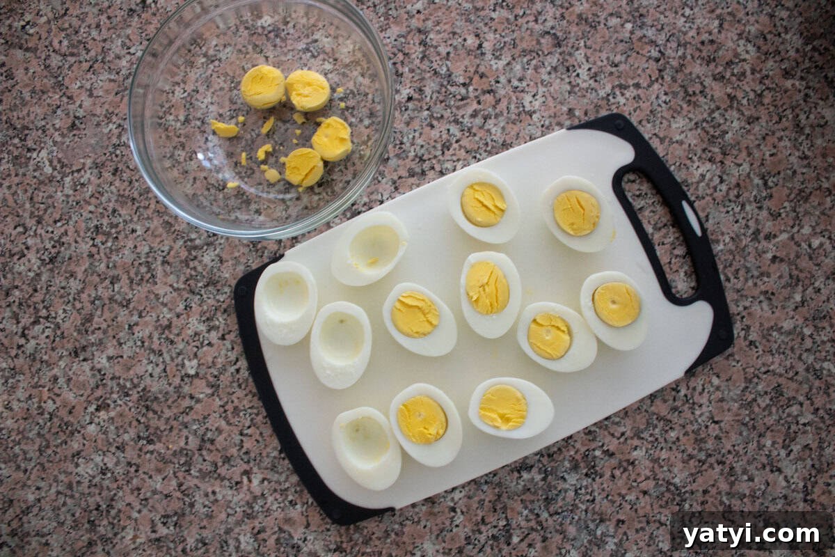 Creamy Egg Salad Without Mayo: Tangy Avocado & Yogurt Recipe 4 Halved hard-boiled eggs with yolks removed are arranged on a white cutting board. A glass bowl containing separated yolks and yolk pieces sits nearby on a speckled countertop.