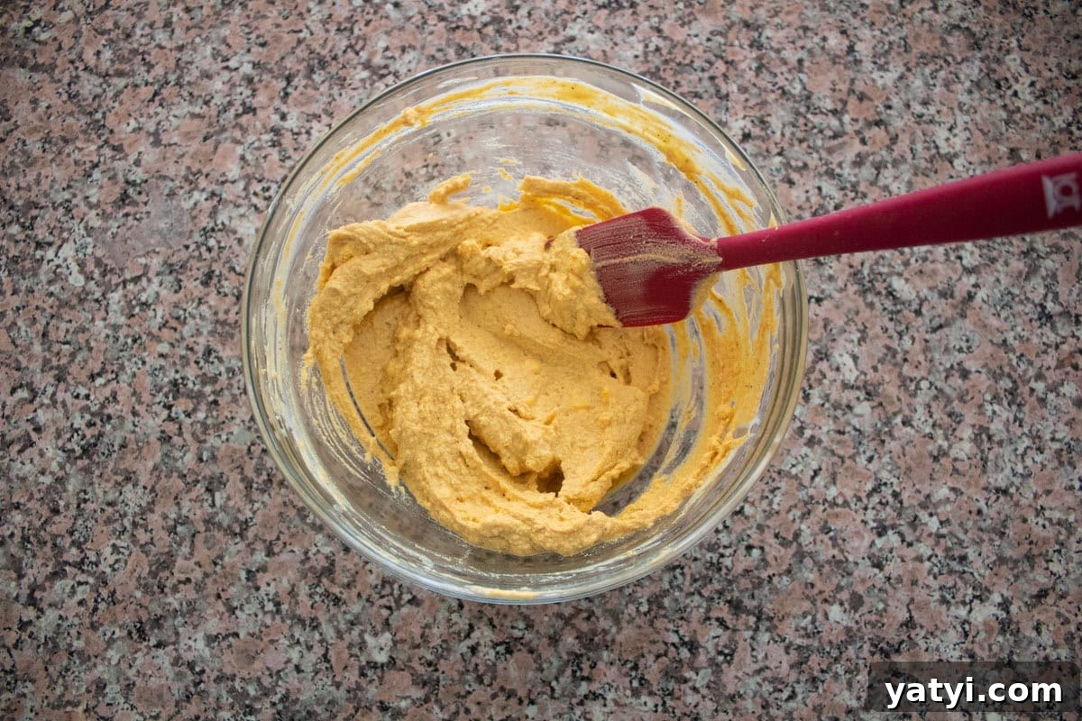 Creamy Egg Salad Without Mayo: Tangy Avocado & Yogurt Recipe 5 A glass bowl with thick, yellowish egg yolk mix being mixed by a red spatula, placed on a speckled countertop.