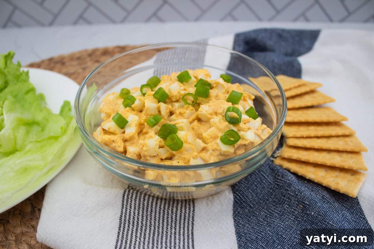 Creamy Egg Salad Without Mayo: Tangy Avocado & Yogurt Recipe 6 A glass bowl of egg salad topped with sliced green onions, placed on a striped cloth beside neatly arranged crackers and a plate of lettuce leaves.