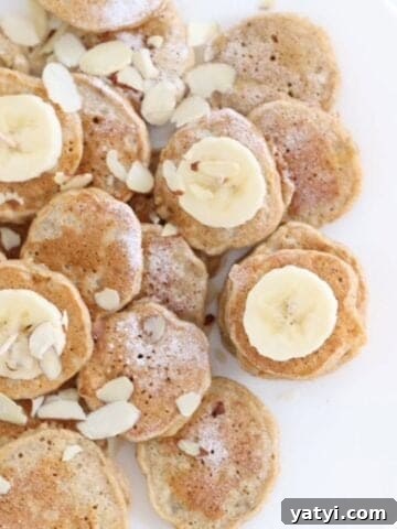 Mini banana pancakes dusted with powdered sugar, topped with sliced banana and almond slices on a white plate.