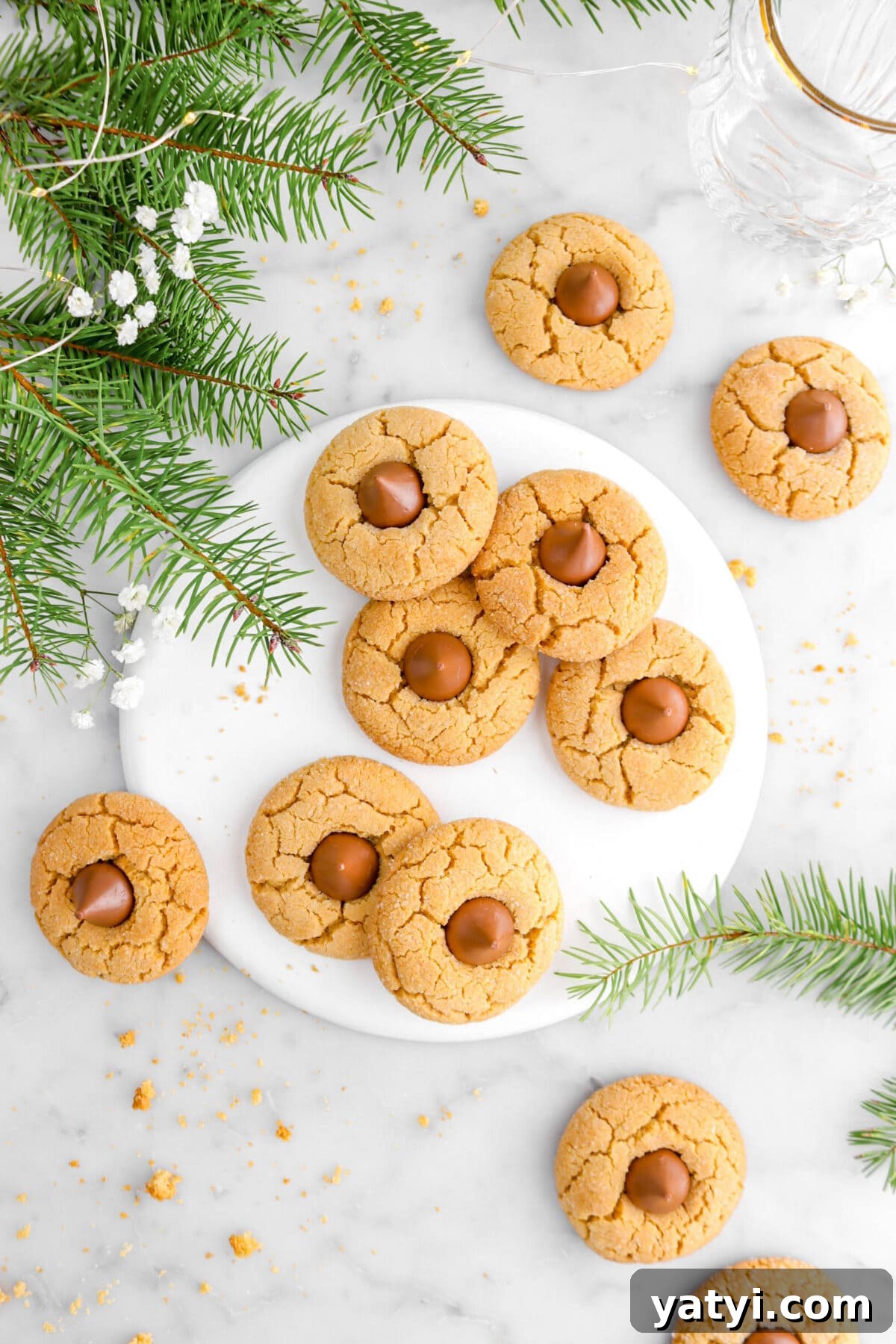 overhead shot of peanut butter blossom cookies with six on white plate, more around on marble surface, with pine greenery and flowers.