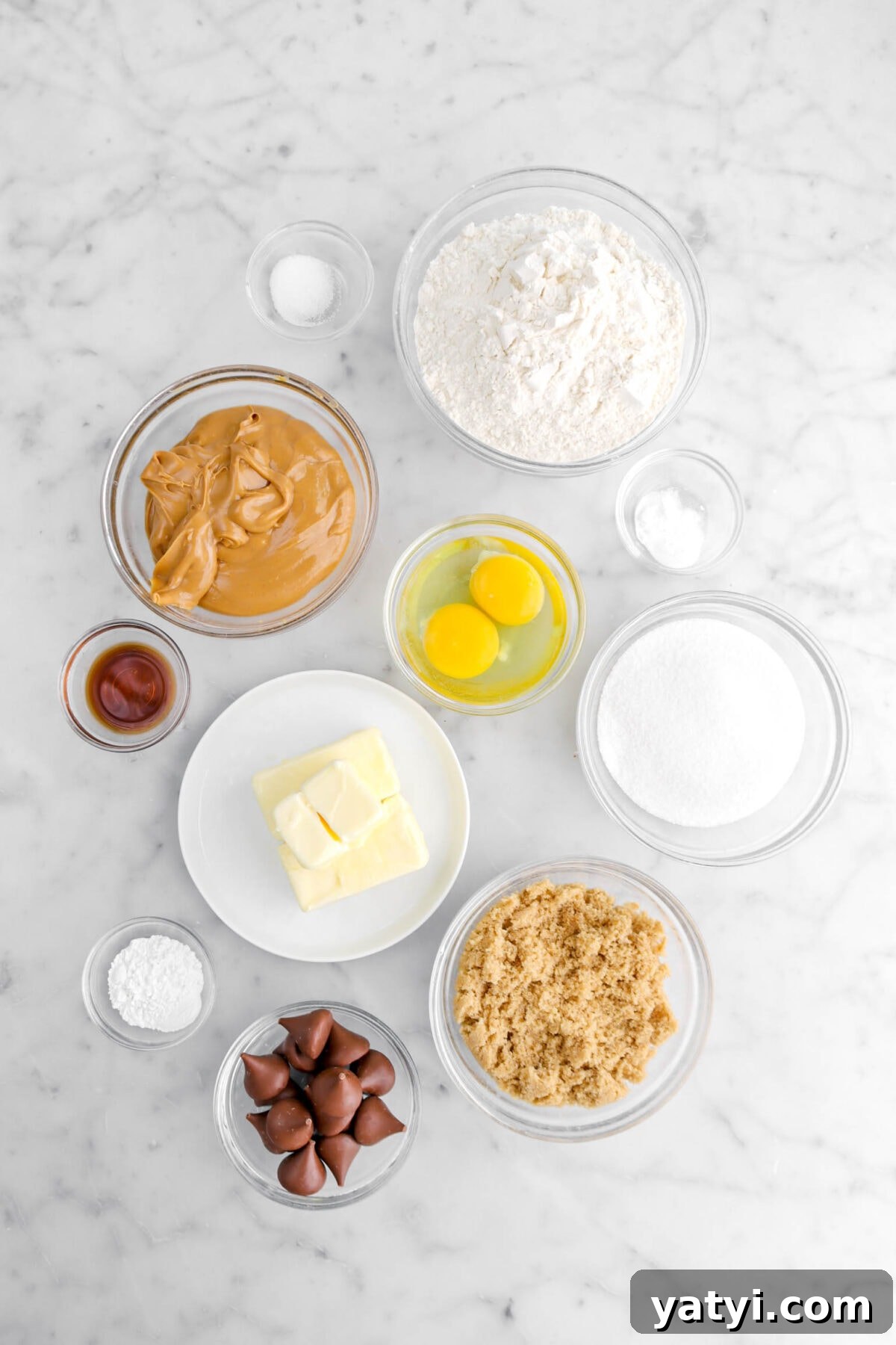 salt, flour, peanut butter, baking soda, eggs, butter, vanilla, baking powder, sugar, brown sugar, and chocolate kisses on marble surface.