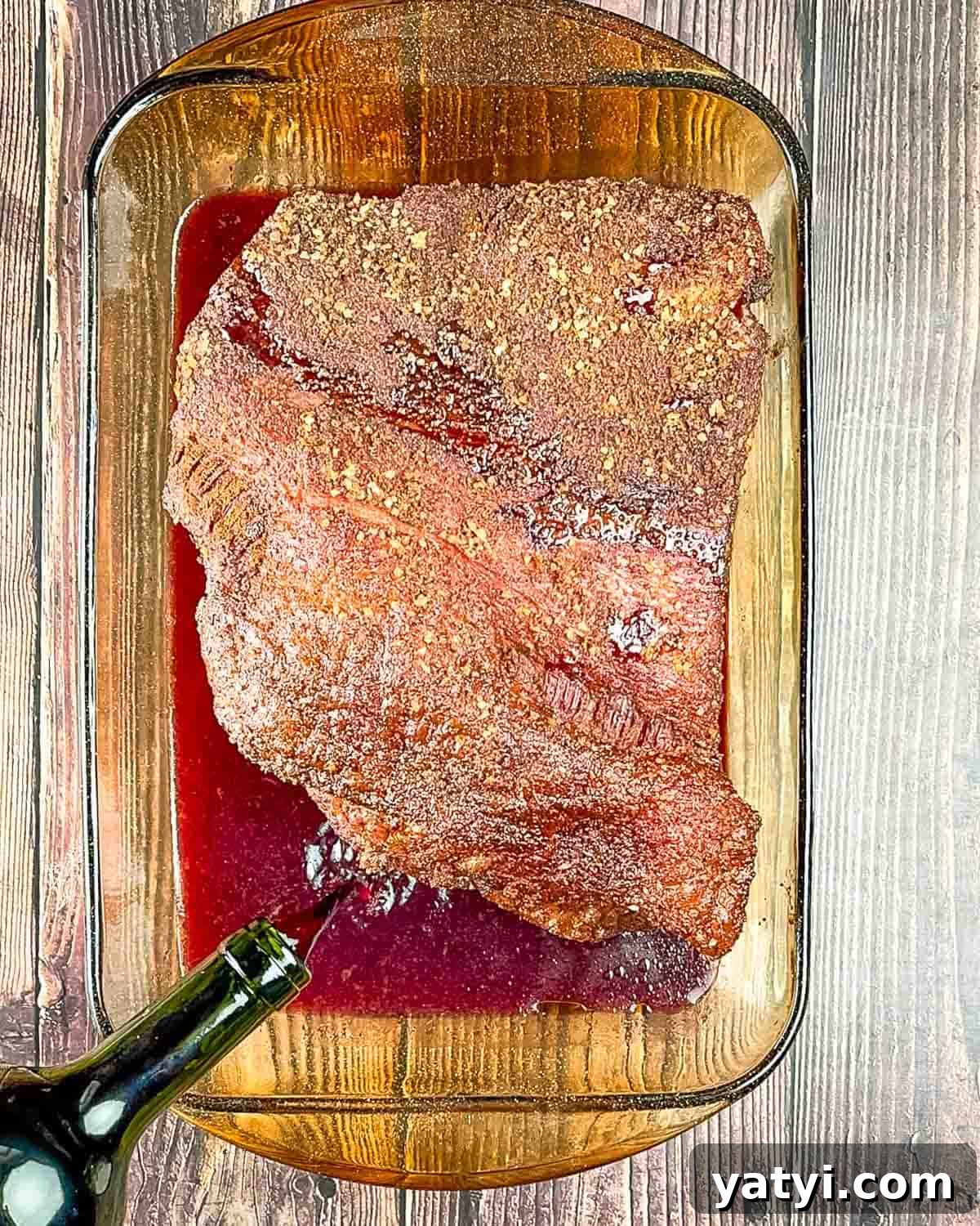 A bottle of red wine being poured into the baking dish with the smoked brisket.
