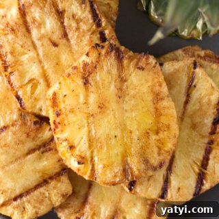 Top down shot of eight grilled pineapple slices on a gray plate with pineapple leaves in the upper right corner.