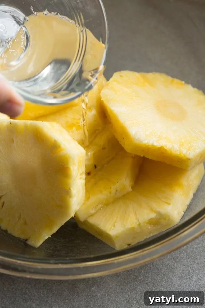 Juicy Grilled Pineapple Recipe — Naturally Sweet, No Added Sugar 7 Pouring coconut oil out of a small clear bowl over pineapple slices in a large clear bowl.
