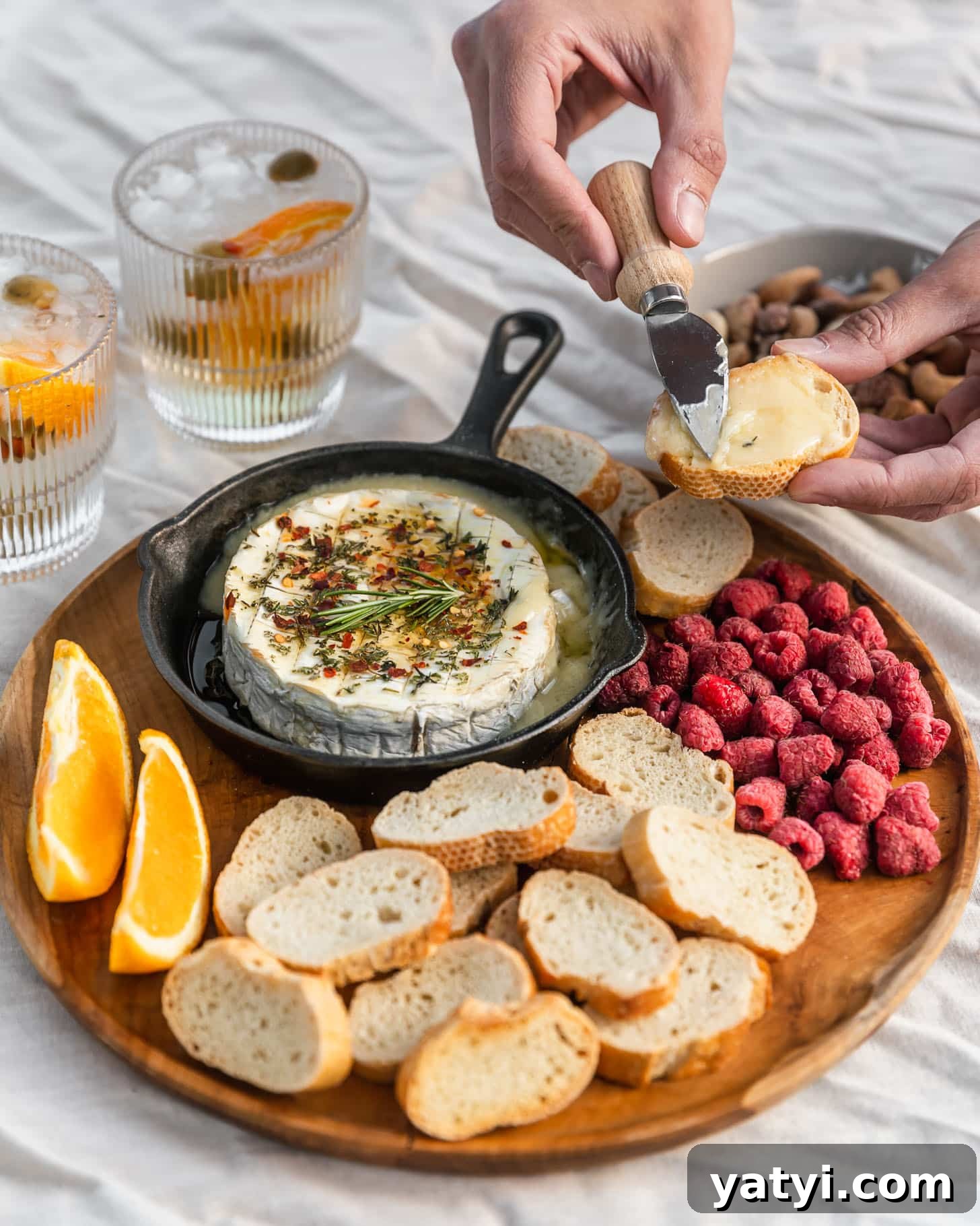 Host an Intimate Dinner Party at Home: Menu, Ambiance & Tips 3 A wood board with baked Camembert, raspberries, oranges, and baguette on a white tablecloth next to a white bowl of nuts and two spritz cocktails.