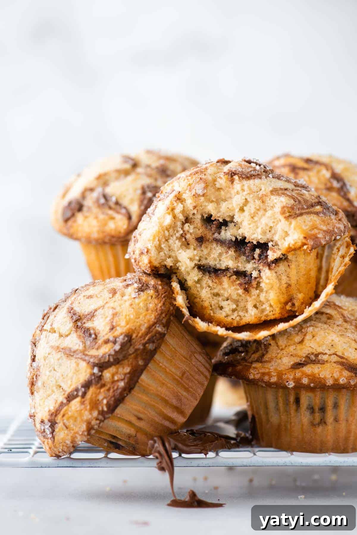 Nutella Swirl Muffins Recipe: Moist Chocolate-Hazelnut Muffins 2 a pile of nutella swirl muffins on a wire rack, with one muffin on its side and the one on top of it with the muffin paper pulled partially off, exposing the layers of nutella inside, and a knife with nutella dripping down from the wire rack
