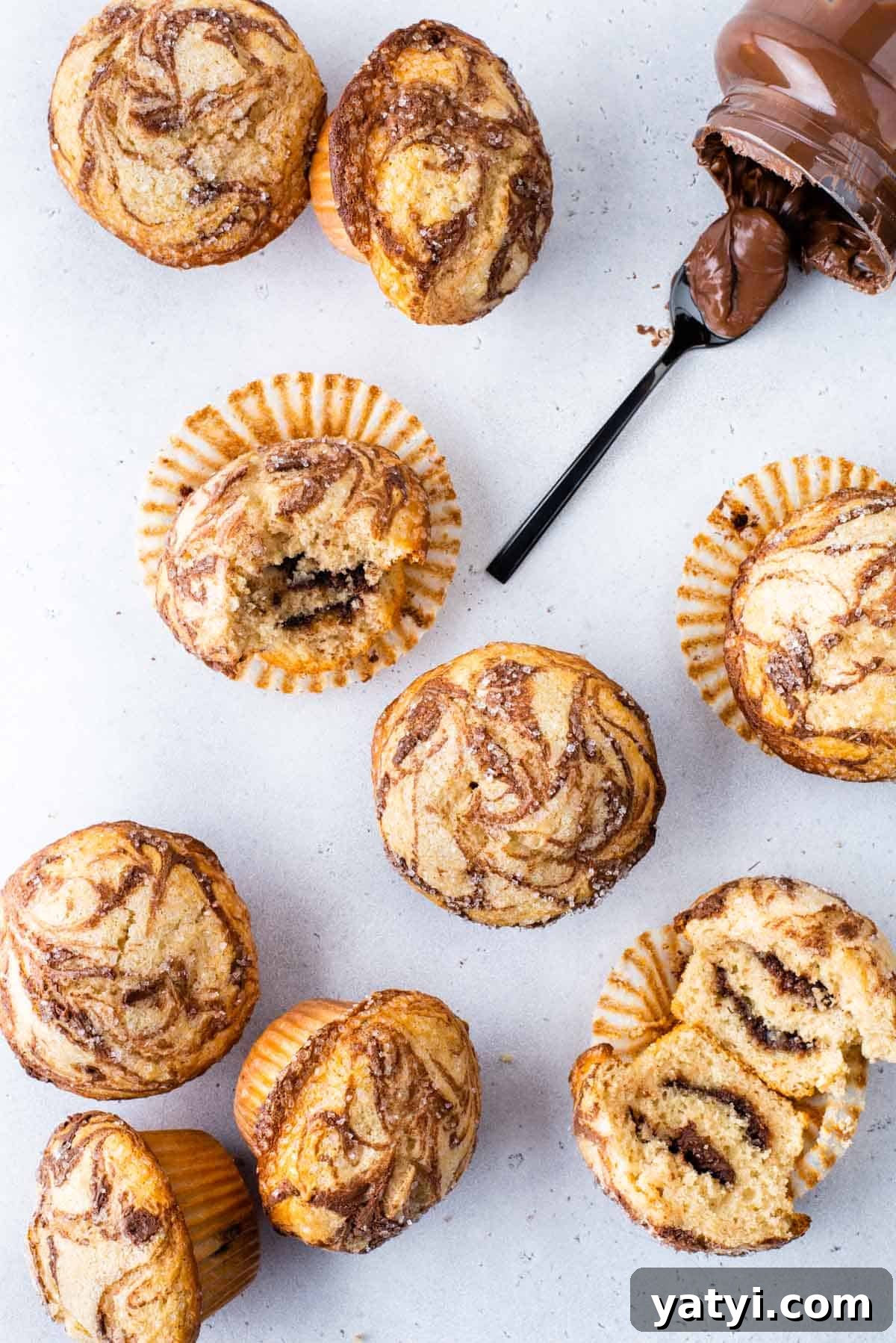 Nutella Swirl Muffins Recipe: Moist Chocolate-Hazelnut Muffins 3 several nutella swirl muffins arranged on a white surface, with some muffins sitting upright, some on their sides, one cut in half exposing the center, and one with a bite out of it, with a nutella jar laying on its side and a spoon of nutella laying beside its opening