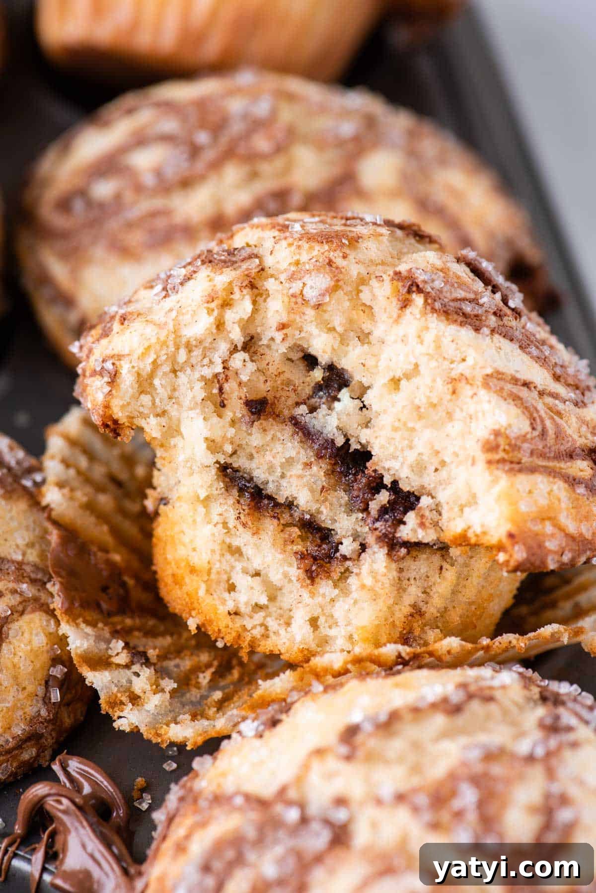Nutella Swirl Muffins Recipe: Moist Chocolate-Hazelnut Muffins 5 a nutella swirl muffin with a bite taken out, exposing the nutella layers inside, resting in its open muffin liner on top of a muffin pan full of more muffins