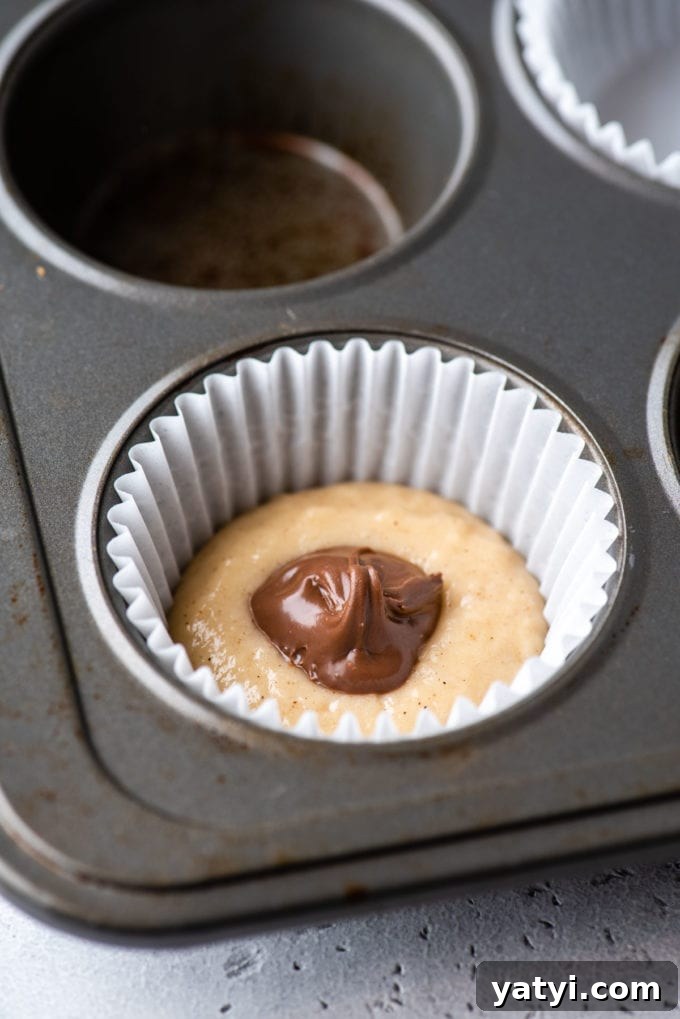 Nutella Swirl Muffins Recipe: Moist Chocolate-Hazelnut Muffins 7 a muffin pan with a white muffin liner in the corner spot and some muffin batter in the bottom of it and a dollop of nutella on top of that